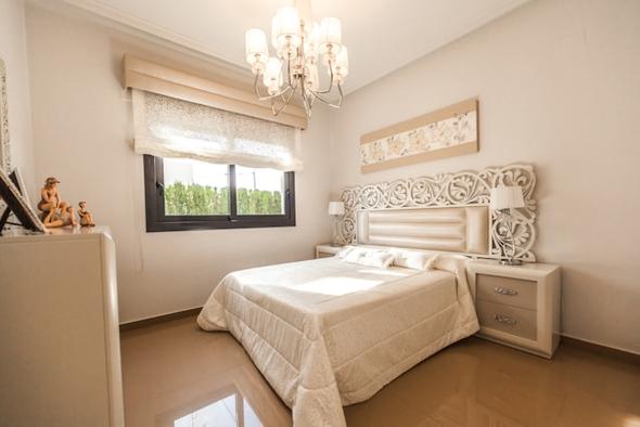 A softly lit, cream-and-beige guest bedroom in a vacation rental property. The room features a queen-sized bed with an ornately carved white headboard, a crystal chandelier, matching nightstands, framed wall art, and a window dressed with a sheer Roman shade — the kind of polished, guest-ready space hosts risk losing payouts on when a double booking forces a cancellation.