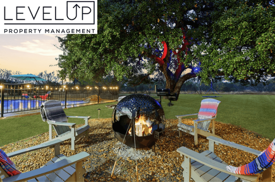 An evening outdoor amenity area at a LevelUp Stays property featuring a glowing spherical metal fire pit surrounded by Adirondack chairs with colorful blankets, set beside a large illuminated oak tree and a fenced swimming pool strung with ambient lights.