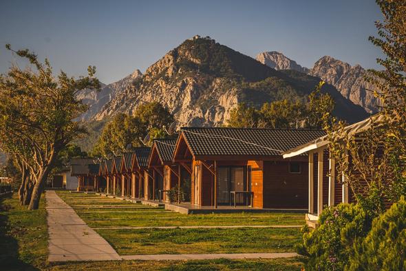 A ground-level photograph of a neatly arranged row of warm-toned wooden cabins with peaked roofs and covered porches, set against a backdrop of towering, rugged mountain peaks. Green grass, mature trees, and shrubs surround the properties, and golden sunset light bathes the scene. This image illustrates the outdoor and nature-focused niche rental category. the type of property that performs well on adventure-travel and glamping-specific booking platforms where guests are searching for mountain retreats, eco-lodges, and remote getaways.