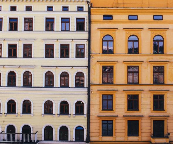 A close-up, symmetrical shot showing the facades of two adjacent multi-story buildings divided right down the middle by a black vertical drainpipe. The building on the left is a soft, pale cream color with neat rows of arched and rectangular dark-wood windows. The building on the right is a warm mustard-yellow color featuring matching classical arched and rectangular window designs. This clean split-screen style composition visually symbolizes a side-by-side comparison, representing the choice between listing on two distinct vacation rental platforms.