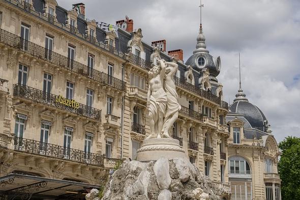 A historic statue in the Place de la Comédie in Montpellier, surrounded by ornate 19th-century buildings.