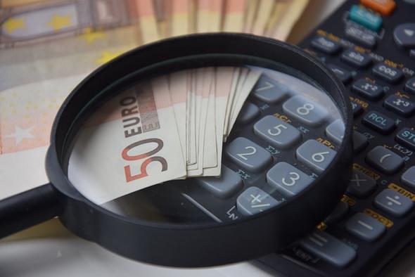 An image depicting rising costs. A magnifying glass focusing on 50 Euro banknotes placed over a calculator, symbolizing the examination of rising costs and financial management.