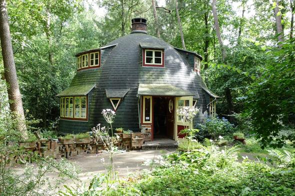 A charming, unconventional dome-shaped house painted dark green with yellow and red window accents sits in a densely wooded clearing. The quirky architecture, chimney, and surrounding overgrown garden with stacked bricks and wildflowers give it a storybook quality. This image represents the unique and unconventional property niche listings that thrive on platforms specializing in unusual stays, tiny homes, treehouses, and architecturally distinctive rentals that attract guests specifically seeking one-of-a-kind experiences they won't find on mainstream booking sites.