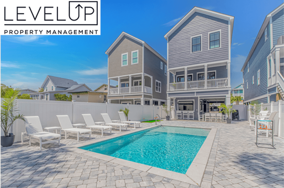 Outdoor patio area of a modern multi-story coastal vacation rental managed by LevelUp Stays, featuring light-toned pavers surrounding a rectangular in-ground pool, white lounge chairs, an outdoor dining space, and a white privacy fence.