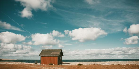 A lone red beach hut sits on a deserted shoreline with no visitors in sight—a visual metaphor for the quiet periods that coastal vacation rental properties experience during the off-season. The vast, empty landscape and open sky emphasize how dramatically demand can drop when peak travel months end, leaving properties waiting for their next booking.