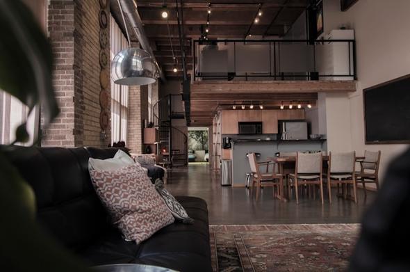 A wide-angle interior view of a spacious, open-concept industrial loft apartment. The design showcases high ceilings with exposed wood beams, metal ductwork, and track lighting. An upper-level mezzanine loft with metal and glass railings overlooks the living area. The main floor features an exposed brick wall, a black leather sofa with patterned cushions in the foreground, an oriental rug, a modern silver dome pendant light, and a wooden dining set. In the background, an open-plan kitchen with warm wood cabinets stands near a spiral staircase leading upstairs