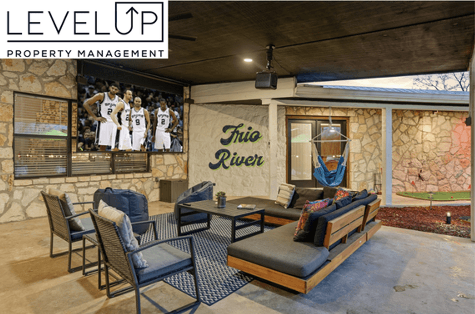 Covered outdoor living and entertainment patio at a LevelUp Stays vacation home, showcasing stone walls, cushioned sectional seating, a hanging hammock chair, "Frio River" wall lettering, and a large projector screen displaying a basketball game.
