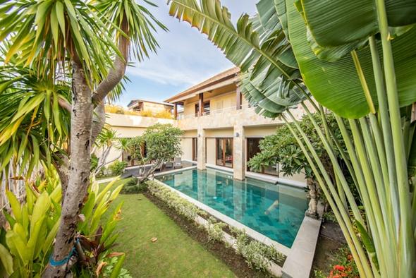 A two-storey vacation rental villa featuring a turquoise private pool, manicured lawn, and tropical landscaping with palm and frangipani trees — showcasing the kind of standout outdoor amenity that attracts more bookings.