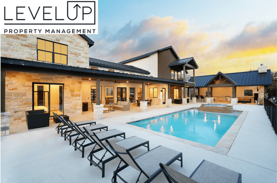 Dusk view of a luxury vacation rental property managed by LevelUp Stays, showcasing a limestone-accented exterior, an illuminated covered back porch, modern poolside lounge chairs, and a lit swimming pool with an integrated hot tub beneath a sunset sky.