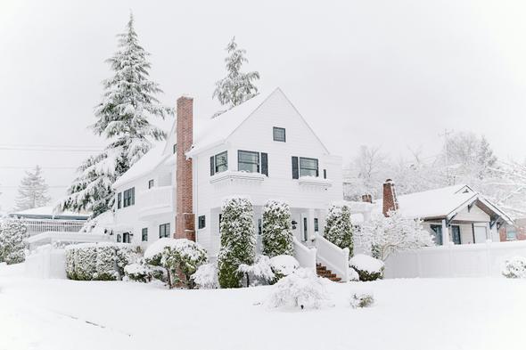 A charming white clapboard home sits quietly under a thick layer of fresh snow, its brick chimney and columned front porch barely visible beneath the winter blanket. Snow-laden evergreen trees and frosted shrubs fill the yard, illustrating the kind of cold-weather slow season that affects vacation rental markets in northern climates during January and February.