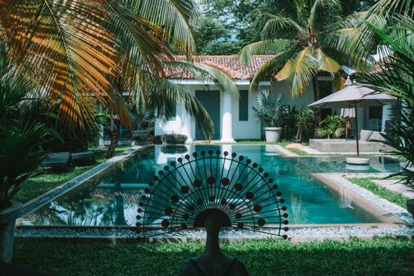 A ground-level view of a tranquil vacation rental property featuring a private swimming pool framed by tall palm trees, a decorative metal sculpture in the foreground, and a patio with a sun umbrella — representing the individual property experience that short-term rental operators manage at scale.